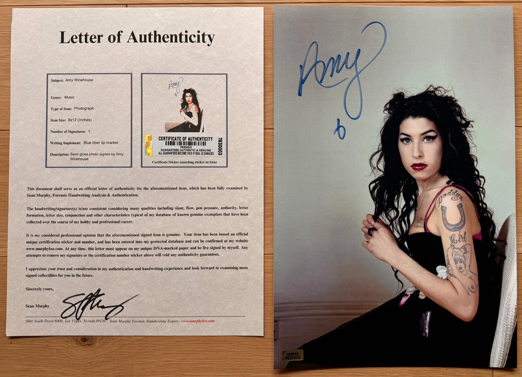An autographed 8x12 photo print of Amy Winehouse, signed in blue ink at the top left. The photo shows her with long dark curly hair, wearing a black top, seated against a light gray wall with COA.