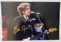 An autographed photo print of Bob Dylan performing on stage, poster signed in gold ink signatures across the lower-left portion. Photograph autographs for sale on 8x12 inch picture.