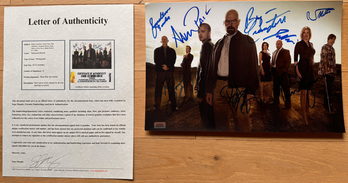 An autographed 8x12 photo print of the cast of Breaking Bad, signed in blue ink across the image, displayed on a wooden surface with COA. RJ Mitte autographs for sale.