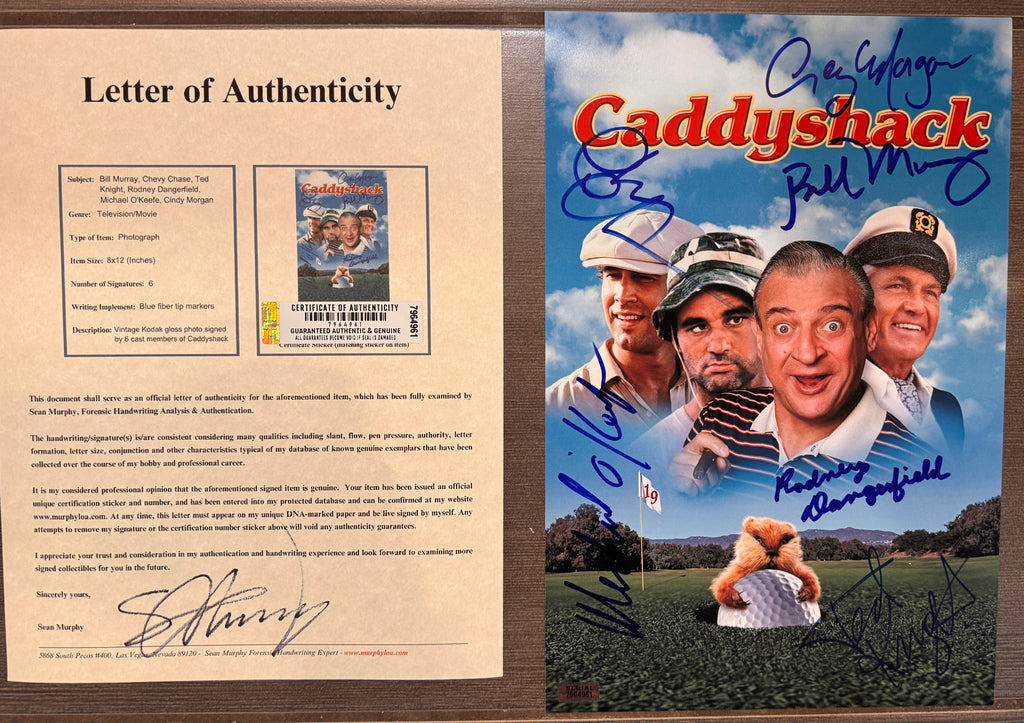 An autographed 8x12 Caddyshack movie photo featuring the cast on a golf course with a teddy bear holding a golf ball. The poster has multiple blue and black ink signatures and is photographed against a brown textured background with COA.