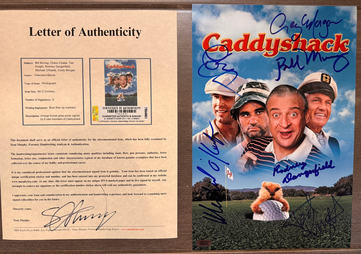 An autographed 8x12 Caddyshack movie photo featuring the cast on a golf course with a teddy bear holding a golf ball. The poster has multiple blue and black ink signatures and is photographed against a brown textured background with COA.