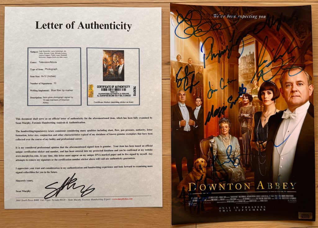 An autographed Downton Abbey movie poster on a light wood background, featuring multiple actor signatures across the front and the film title at the bottom with COA.