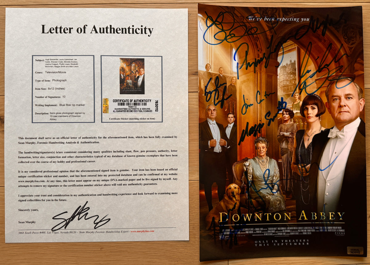 An autographed Downton Abbey movie poster on a light wood background, featuring multiple actor signatures across the front and the film title at the bottom with COA.