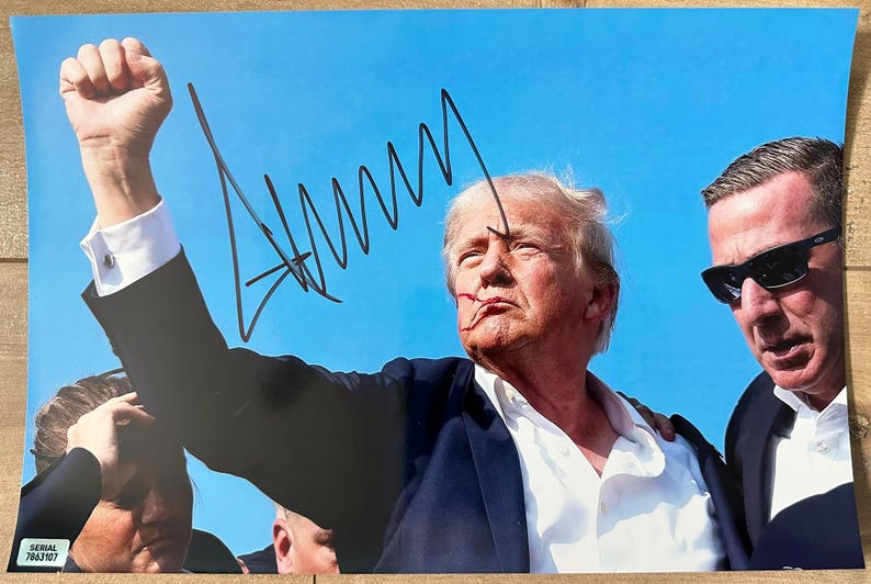 An autographed 8x12 photograph of Donald Trump raising his fist during his attempted assassination on July 13, 2024 in  Butler, Pennsylvania. Signed photo in black ink markers signature. Poster autographs for sale on 8x12 inch picture.