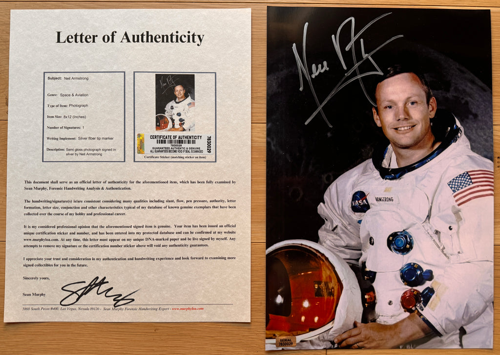 An autographed 8x12 photo print of astronaut Neil Armstrong, signed in silver marker across the top left. The print shows Armstrong in his white NASA spacesuit holding a helmet, set against a dark space background, and is photographed flat on a light wood surface with COA.
