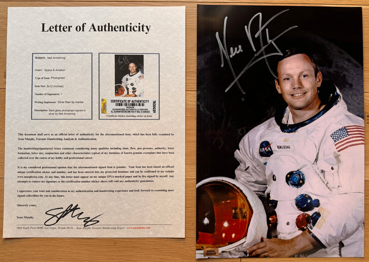 An autographed 8x12 photo print of astronaut Neil Armstrong, signed in silver marker across the top left. The print shows Armstrong in his white NASA spacesuit holding a helmet, set against a dark space background, and is photographed flat on a light wood surface with COA.