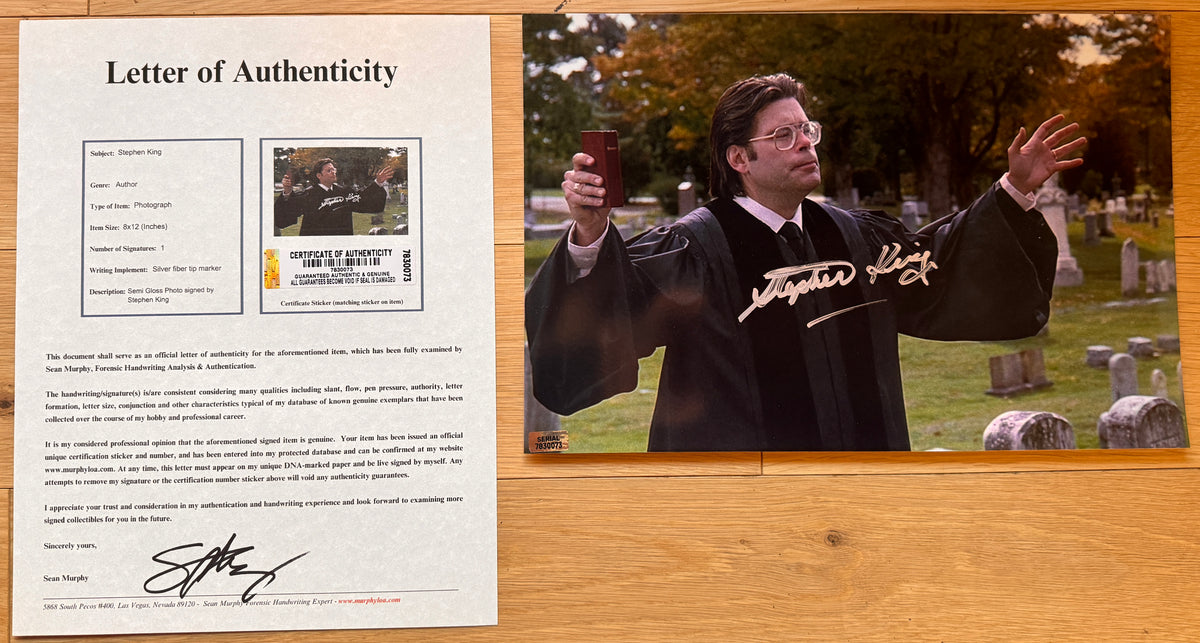 A signed photograph of actor Stephen King in a black graduation gown and tie, holding a phone in a cemetery. The photo features a white script signature across the chest and is framed by a light wood border with COA.