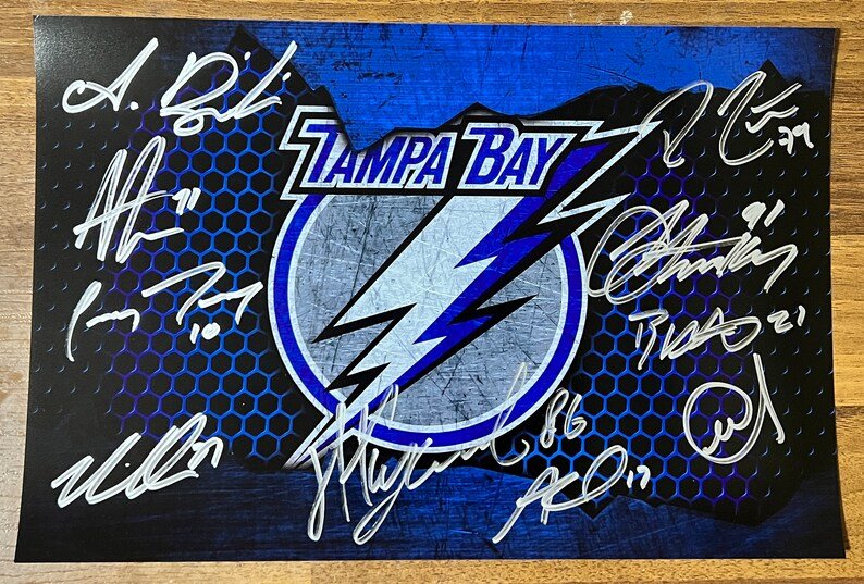 A team signed Tampa Bay Lightning hockey poster. Signed photo of Steven Stamkos, Victor Hedman, Nikita Kucherov, Alex Killorn, Brayden Point, Andrei Vasilevskiy, Anthony Cirelli, Corey Perry, Ross Colton and Mikhail Sergachev in silver ink markers. Photograph autographs for sale on 8x12 inch picture.