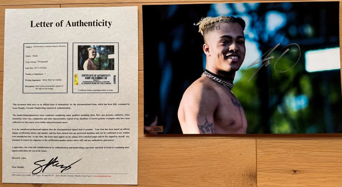 An autographed photo print of rapper XXXTentacion, signed in silver marker across the right side, displayed on a light wood surface. The photo features a smiling XXXTentacion with blonde-tipped hair and tattoos, wearing a silver chain necklace against a dark, blurred outdoor background. autograph on an 8x12 inch photograph poster for sale with COA.